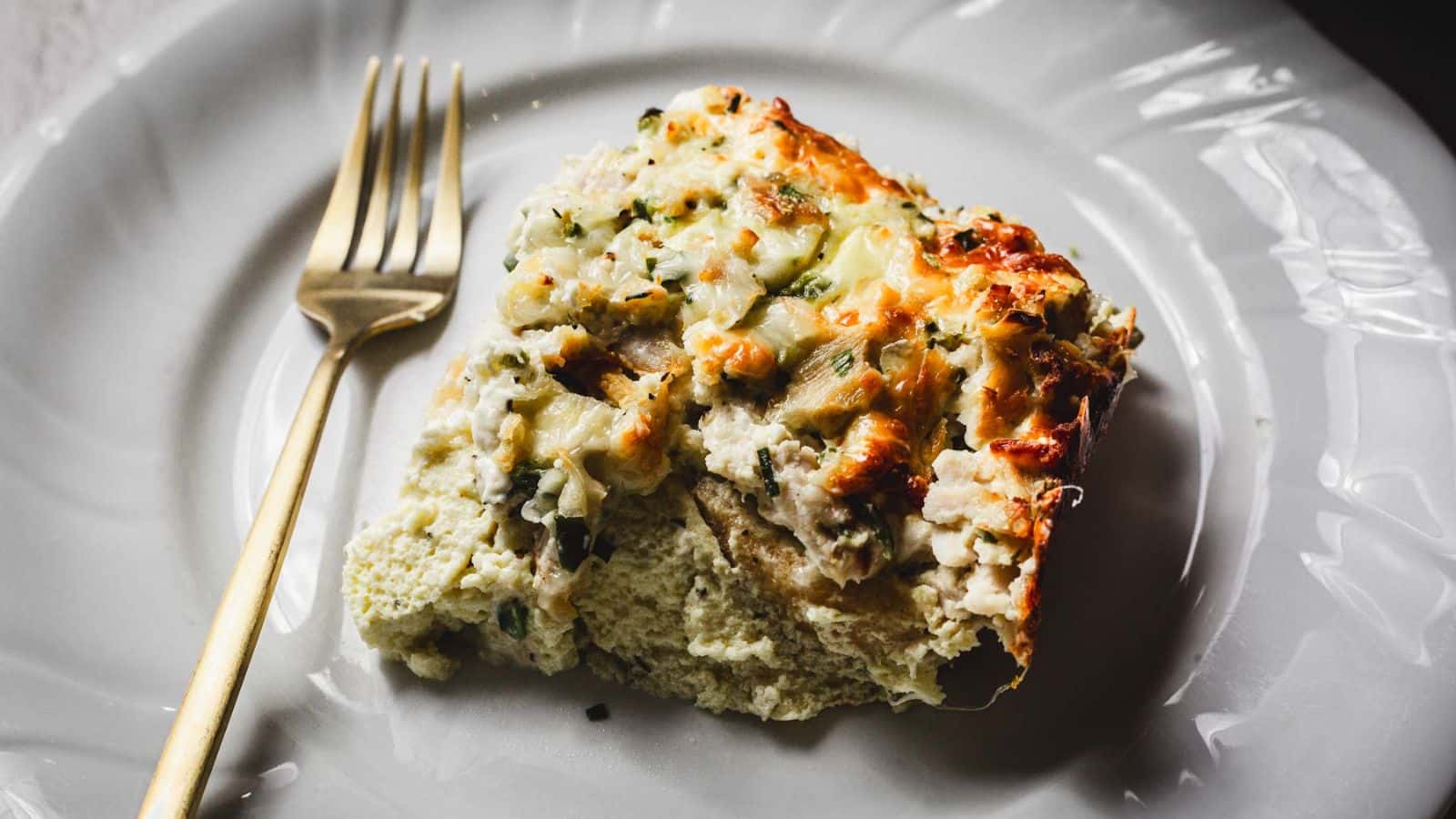 A person is using a spatula to serve a portion of baked mashed potato casserole from a metal baking dish. The casserole is topped with melted cheese and sprinkled with herbs.
