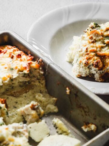 A baking dish filled with a baked casserole topped with melted cheese. A portion is served on a white plate beside it. The casserole appears creamy and slightly browned on top.