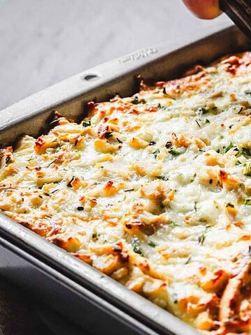 A person is using a spatula to serve a portion of baked mashed potato casserole from a metal baking dish. The casserole is topped with melted cheese and sprinkled with herbs.