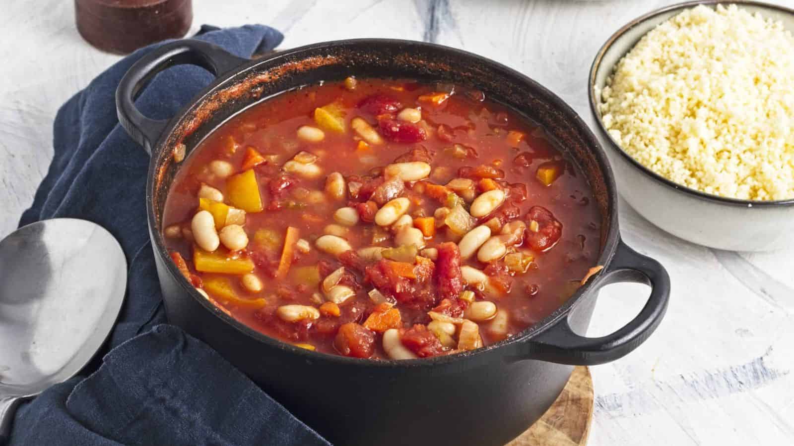 A black pot filled with a tomato-based stew containing white beans, bell peppers, and other vegetables. The pot rests on a dark cloth with a silver spoon nearby. A bowl of couscous sits beside the pot on a white surface.
