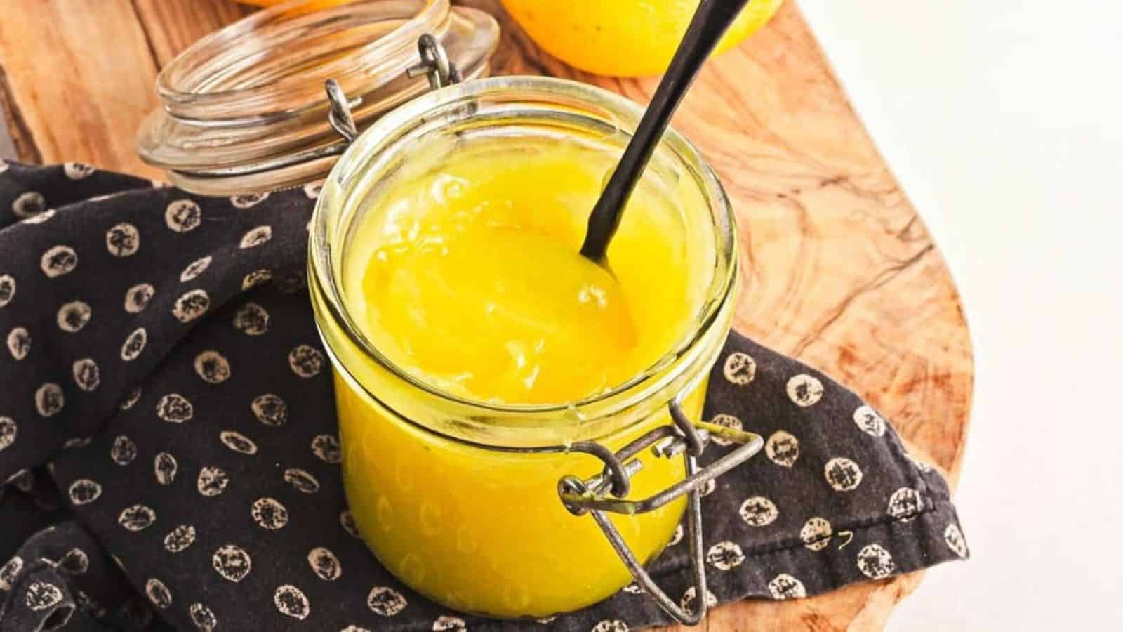 A glass jar filled with yellow lemon curd sits on a wooden surface. A black spoon is inserted into the jar. The jar is resting on a black and white patterned cloth. A partially visible lemon is in the background.