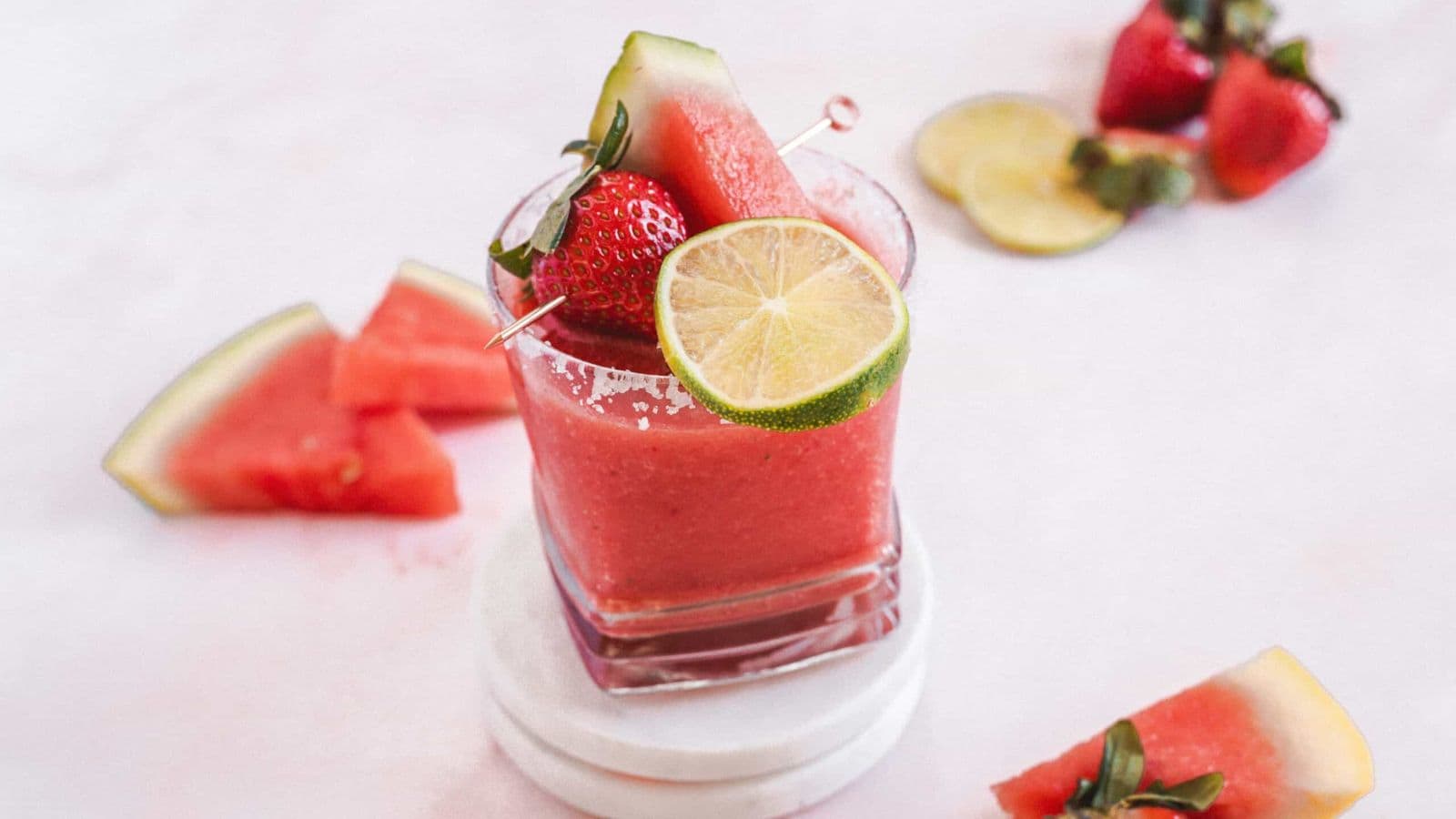 A glass of pink smoothie garnished with a watermelon slice, lime wheel, and strawberry on a skewer stands on white coasters. Surrounding it are strawberries, watermelon slices, and a lime slice on a white surface.