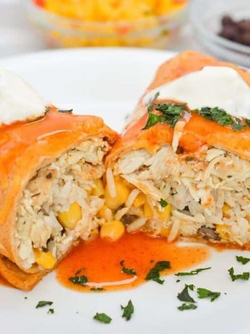 A burrito cut in half on a white plate, topped with creamy sauce and garnished with cilantro, makes for one of those perfect family meals. The filling includes shredded meat, corn, and rice. In the background, there are small bowls of shredded cheese and black beans.