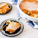 Two servings of sweet potato soufflé on patterned black plates, each with a gold spoon. The light and fluffy soufflé is sprinkled with powdered sugar. A larger dish of the dessert is nearby, partially served, on a white and blue cloth.