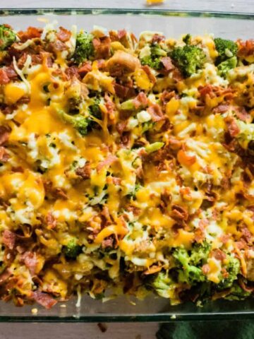 A rectangular glass baking dish filled with a casserole topped with melted cheese. Visible ingredients include pieces of broccoli, bacon, and turkey, with a browned top layer. The dish rests on a wooden surface near a green cloth.