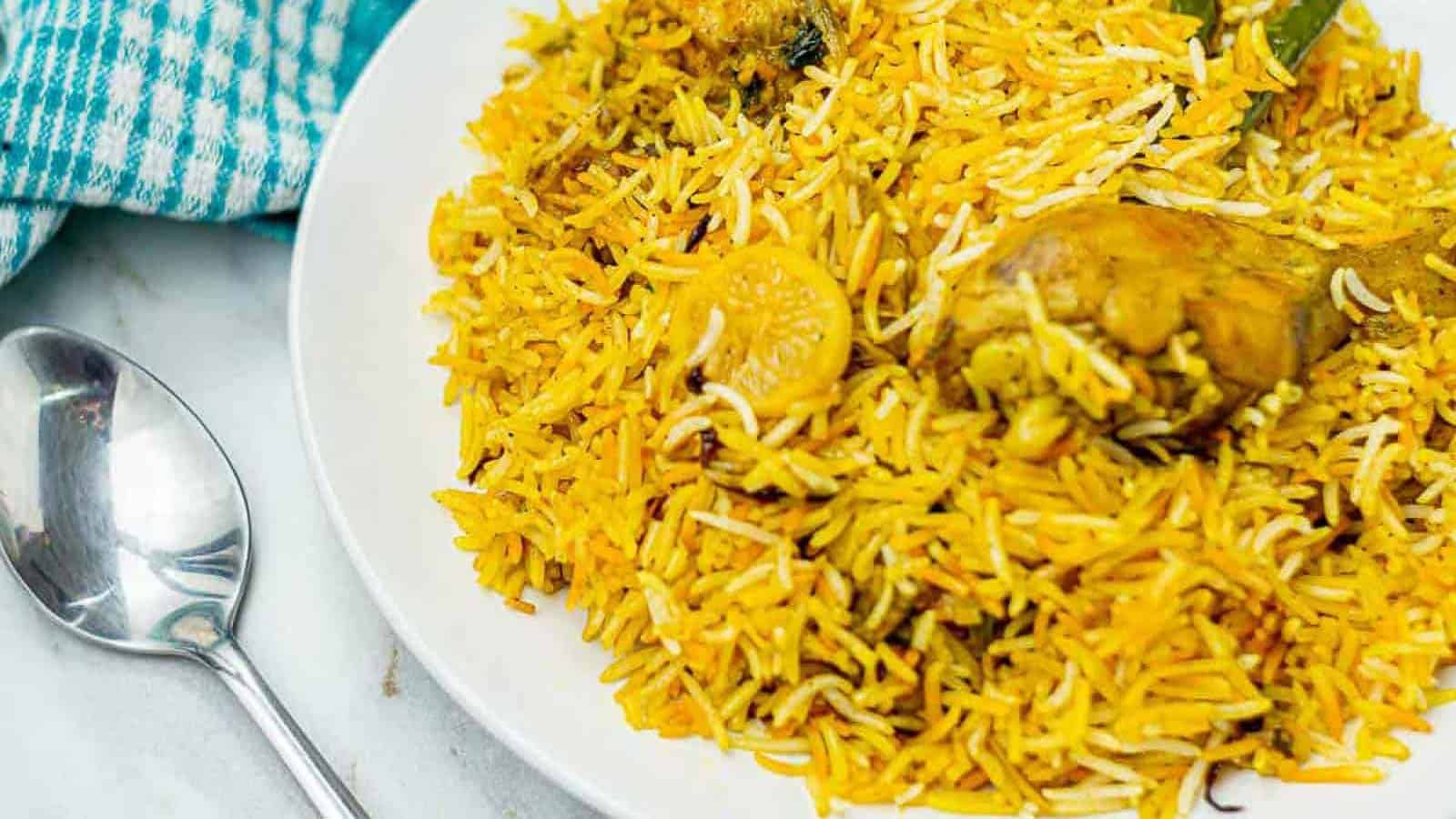 A plate of biryani with yellow and white basmati rice, chunks of chicken, and visible spices. A spoon is placed next to the plate on a marble surface, with a blue and white checkered cloth nearby.