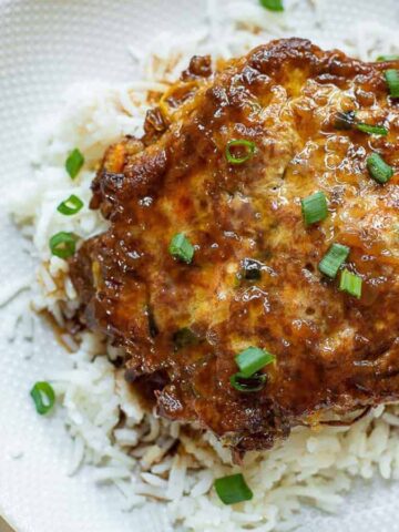 A dish featuring a brown grilled patty on top of white rice, garnished with chopped green onions. The food is presented on a white, textured plate with a cloth napkin partially visible in the background.