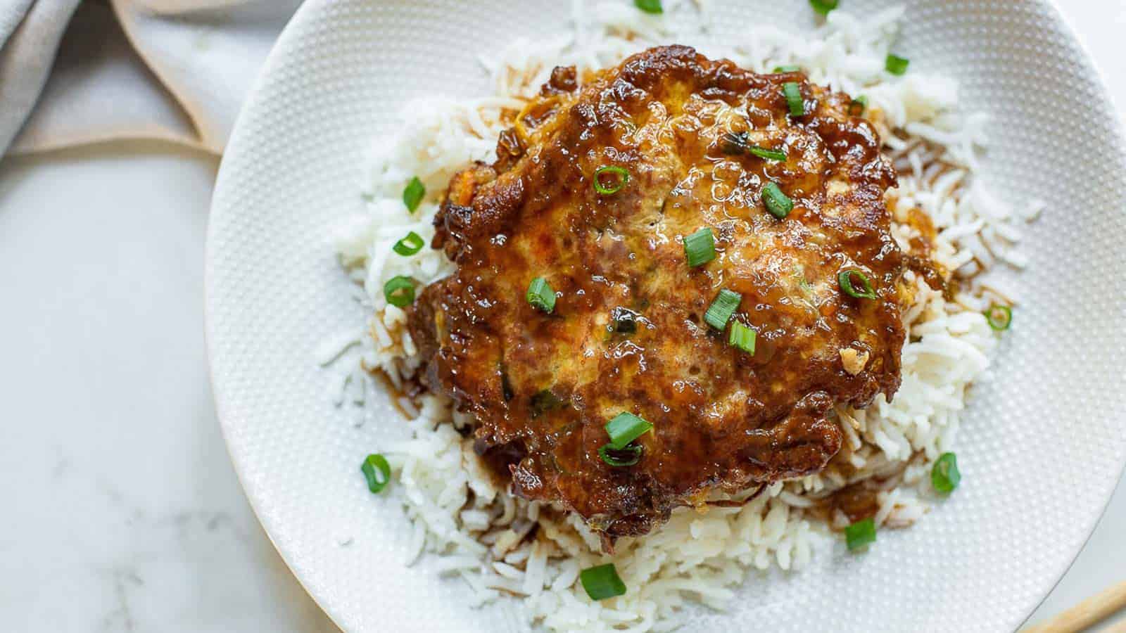 A dish featuring a brown grilled patty on top of white rice, garnished with chopped green onions. The food is presented on a white, textured plate with a cloth napkin partially visible in the background.