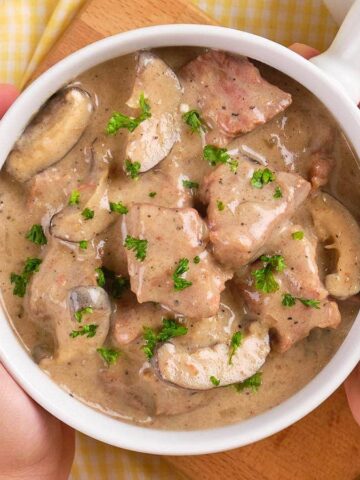 A person holds a bowl of creamy beef stroganoff garnished with parsley. The dish includes sliced mushrooms and beef pieces. Nearby, there is a bowl of quinoa, a fork, and a spoon on a yellow checkered tablecloth.