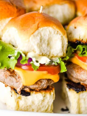 Mini cheeseburgers on a plate with toasted buns, lettuce, tomato, red onion, cheddar cheese, and burger patties. Toothpicks hold them together.
