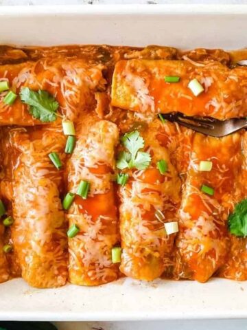 A white rectangular dish brimming with cheesy enchiladas is adorned with melted cheese, chopped green onions, and cilantro. Two metal forks invite you to dive into this main dish. A green cloth and a lime wedge peek in from the side, enhancing the perfect recipe setting.