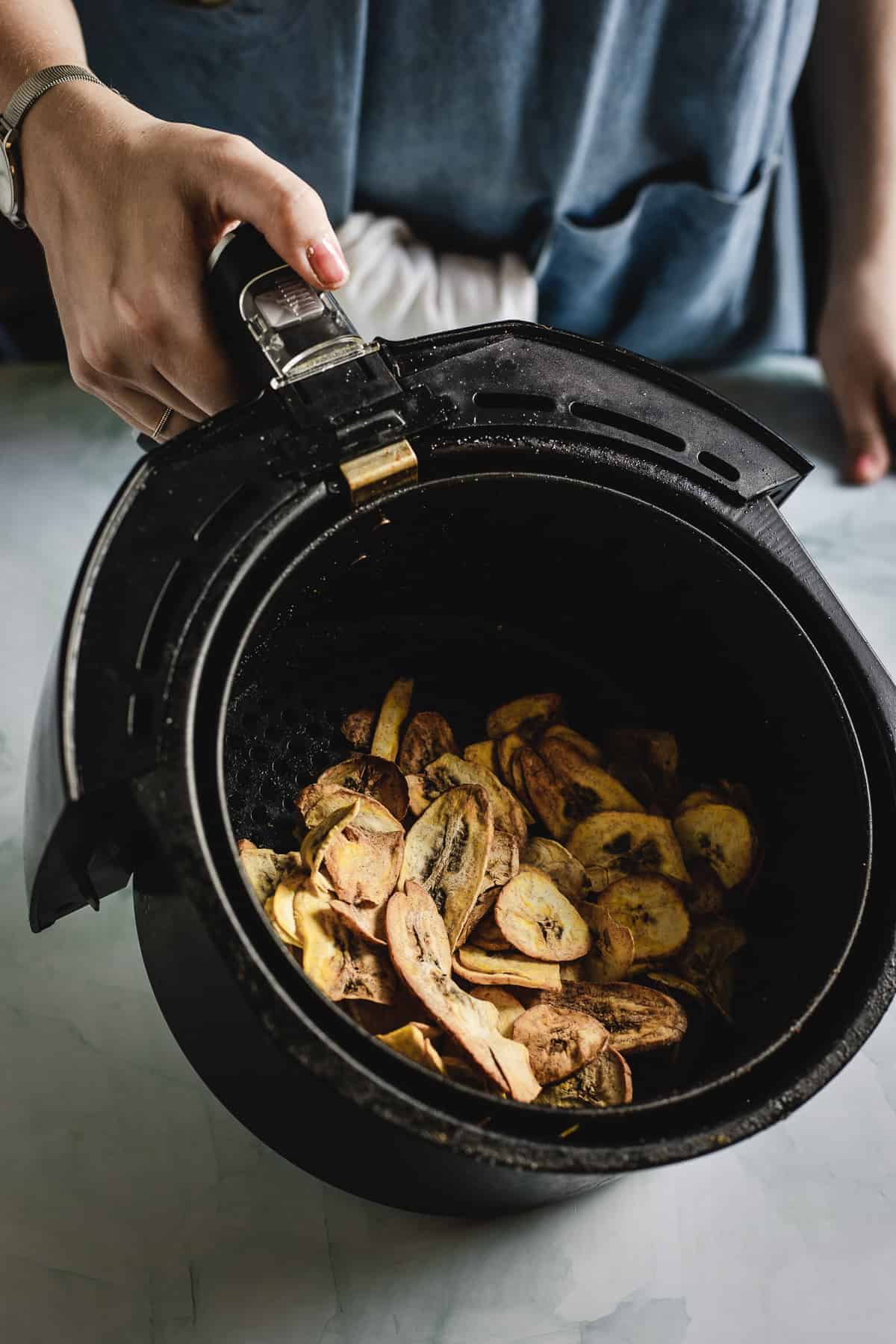A person holds the basket of an air fryer, an essential guide to modern cooking, containing slices of cooked plantains. The sleek black air fryer with a handle complements their blue top, their hand gracefully near the handle.