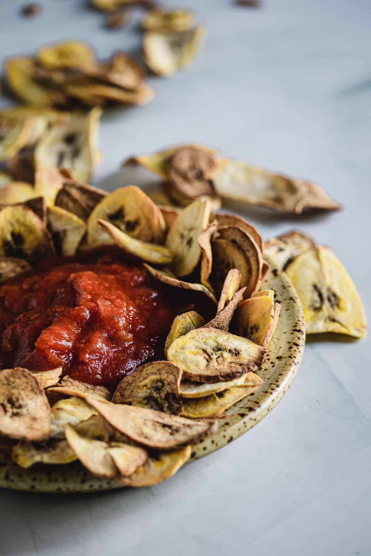 A plate filled with banana chips arranged in a circle around a serving of red salsa on a light surface, with additional banana chips scattered in the background.