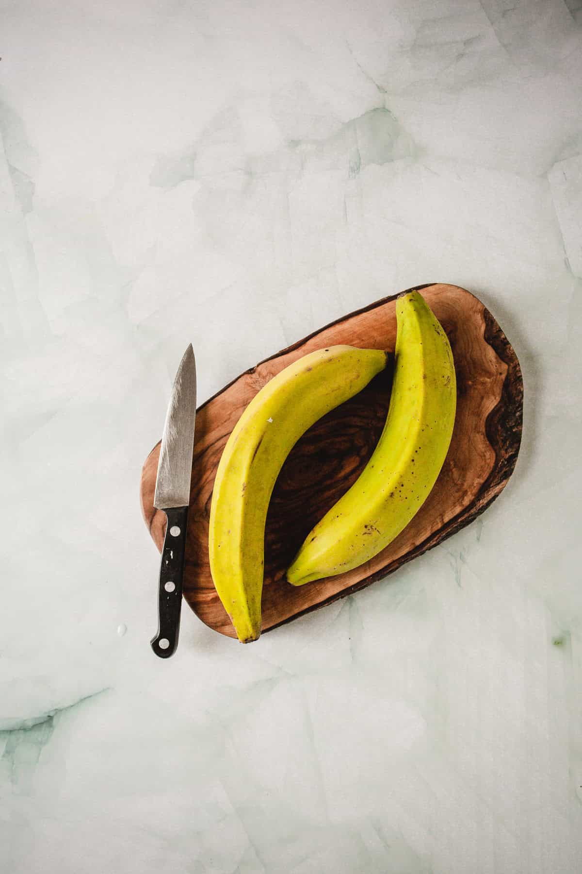 A wooden cutting board with two unpeeled plantains and a kitchen knife is placed on a light marble surface.
