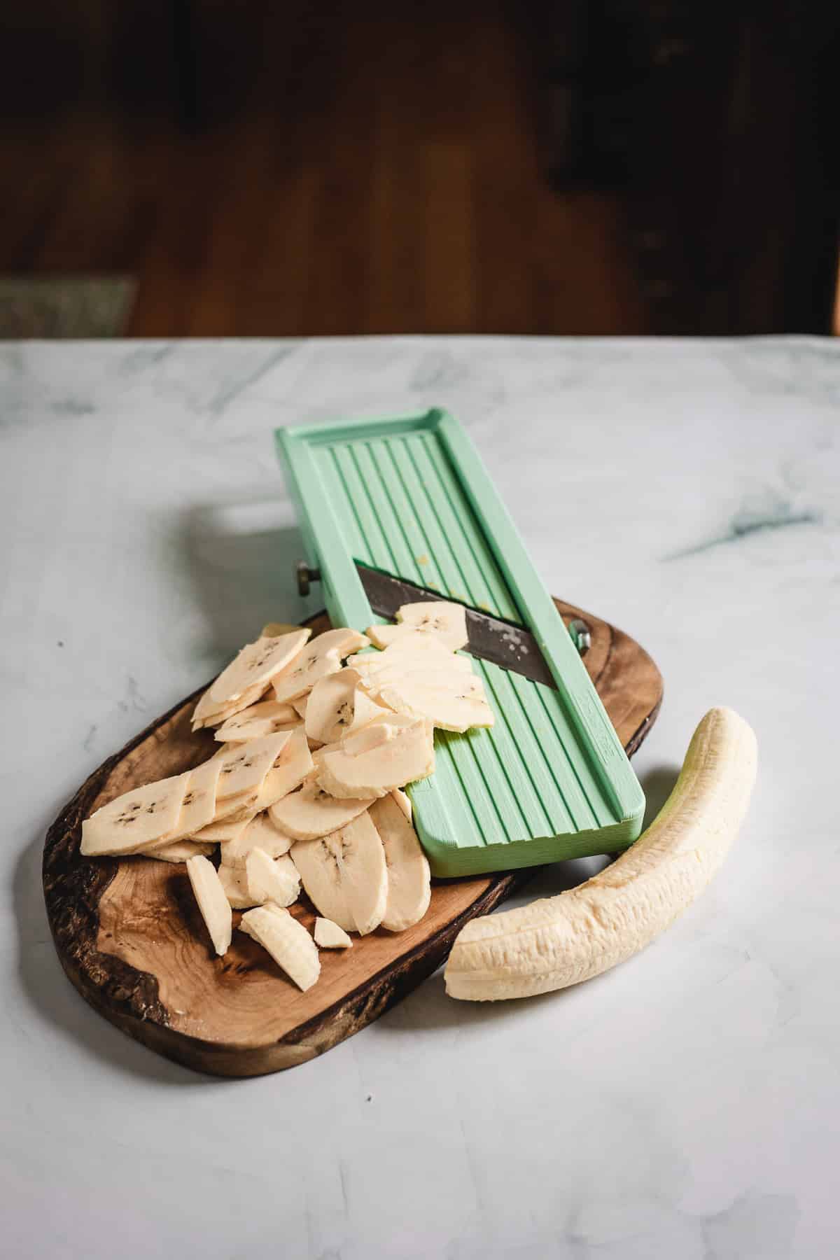 A wooden board with thin banana slices, a whole peeled banana, and a green mandoline slicer on a white surface.