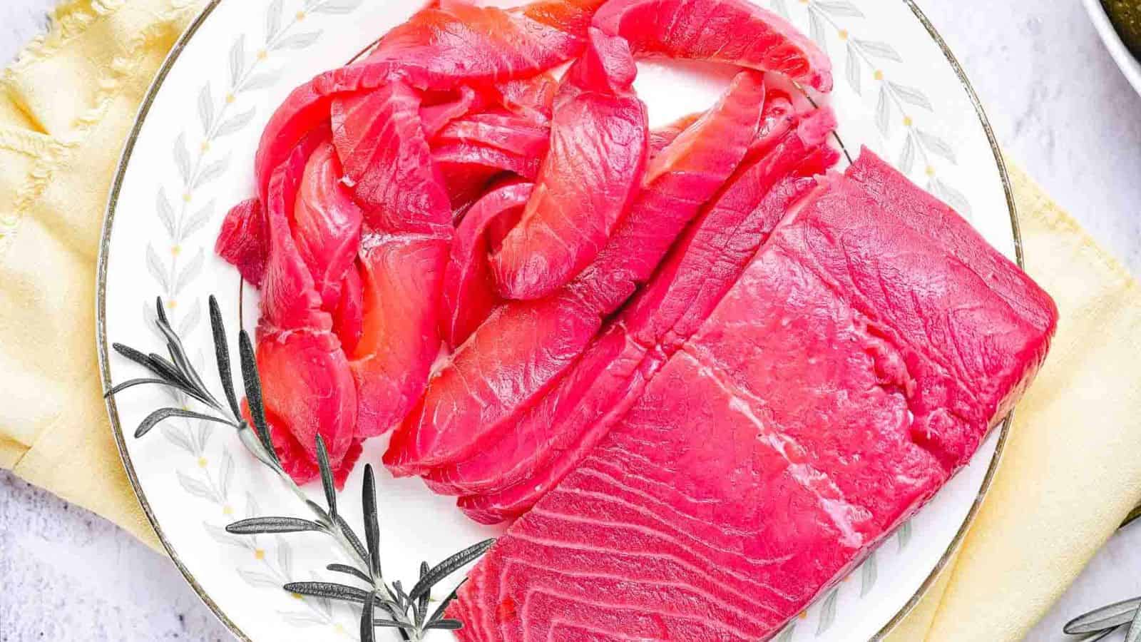 A white plate with a gold rim holds several slices and a block of raw, bright pink tuna. A sprig of rosemary lies on the left side of the plate, which rests on a pale yellow cloth.