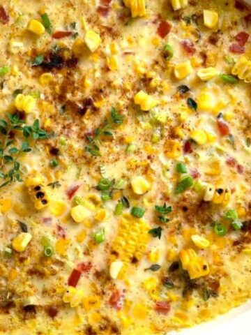 A baked corn casserole in a round dish, topped with grilled corn kernels, chopped green herbs, and red peppers. The surface is browned and slightly crispy, resting on a wooden surface next to a striped cloth.
