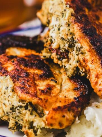 Two pieces of stuffed and grilled chicken breast with a browned, crispy exterior are served on a plate next to a portion of mashed potatoes. The chicken stuffing appears to contain herbs and cheese.
