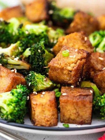 A plate of stir-fried tofu and broccoli topped with sesame seeds makes a delightful kid-friendly entree. Served on a white plate with a fork on the side, this nutritious meal stretches your grocery budget while featuring a red checkered napkin partially visible in the backdrop.