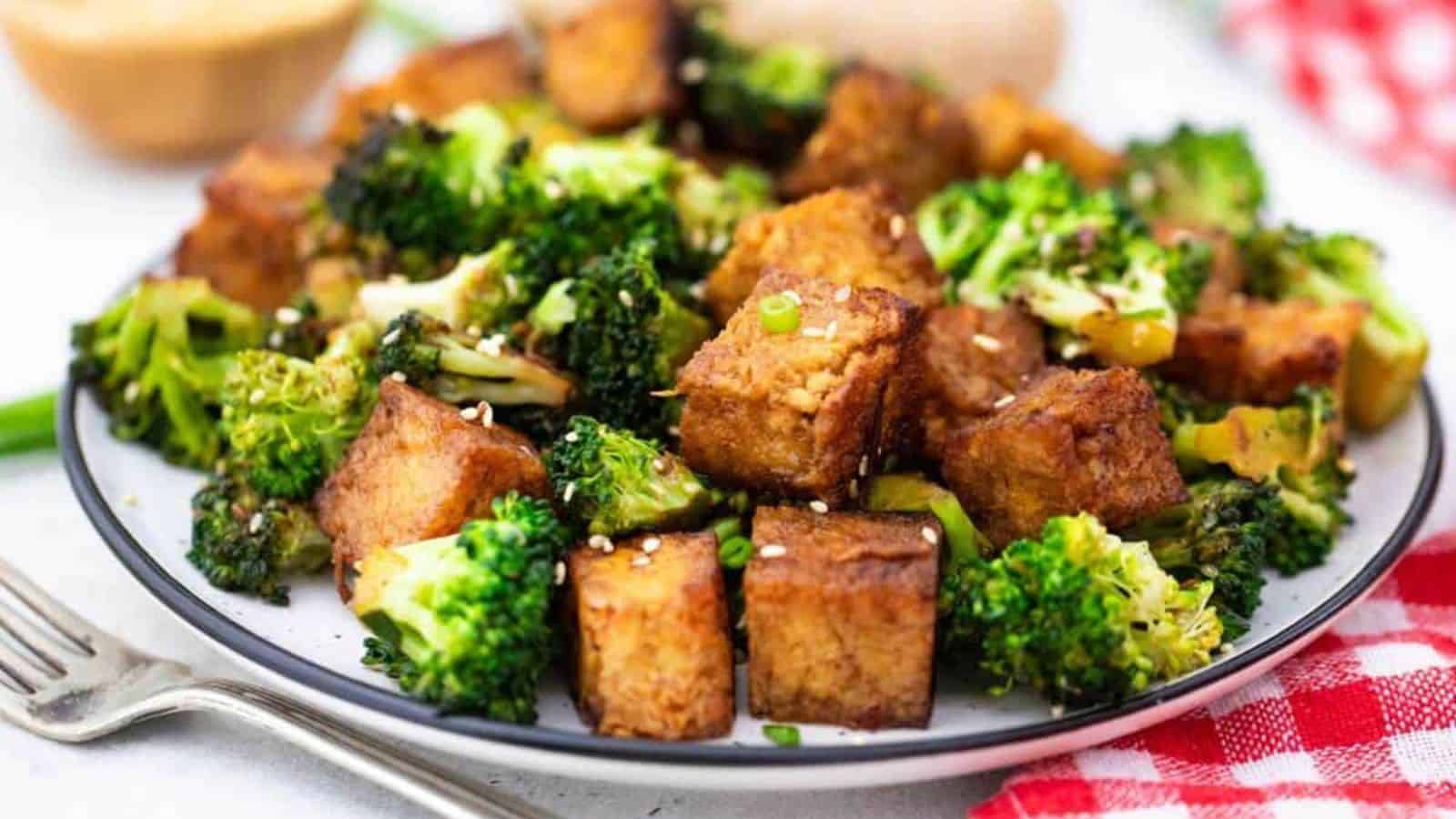 A plate of stir-fried tofu and broccoli topped with sesame seeds makes a delightful kid-friendly entree. Served on a white plate with a fork on the side, this nutritious meal stretches your grocery budget while featuring a red checkered napkin partially visible in the backdrop.