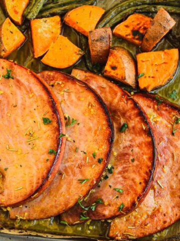 Sliced ham alongside roasted sweet potatoes and green beans on a baking tray. A sprig of rosemary and a small bottle of honey are visible nearby.