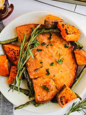 A white plate with a serving of glazed ham garnished with rosemary, surrounded by roasted sweet potato chunks and green beans. A gray napkin and a bottle of syrup are beside the plate.