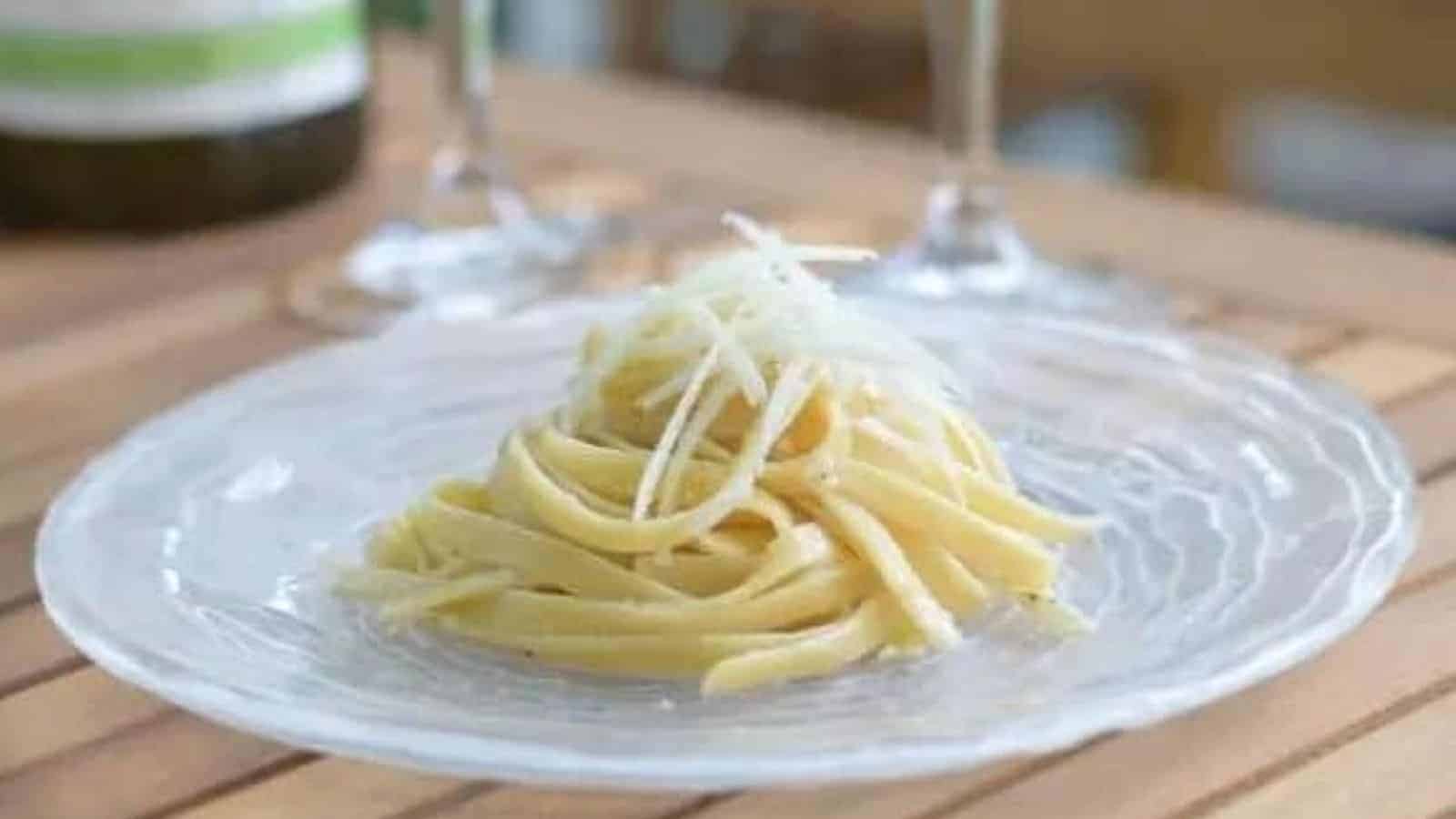 A clear glass plate holds a serving of pasta topped with grated cheese, perfect as a rewarding meal. The background, complete with two wine glasses and the neck of a wine bottle, sets a cozy scene on the wooden surface&mdash;ideal for getting through the week or indulging in weekend recipes.