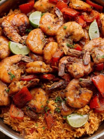 A skillet filled with cooked shrimp, red bell peppers, and lime wedges served over seasoned rice. The skillet sits on a woven placemat next to a beige napkin.