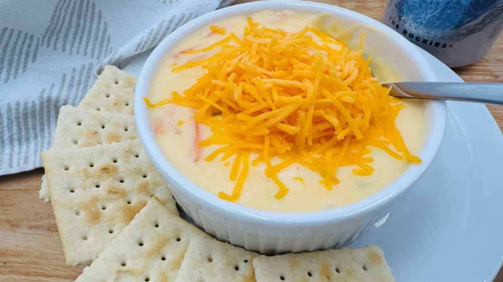 A white bowl of creamy soup, reminiscent of Grandma recipes, is topped with shredded cheddar cheese. The bowl sits on a plate surrounded by square saltine crackers. A spoon rests inside, while a gray-striped cloth provides a cozy backdrop, evoking the warmth of family meals.