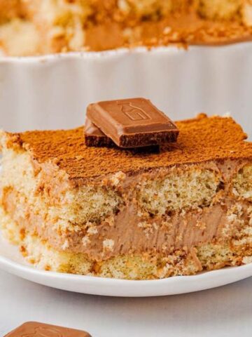 A slice of chocolate tiramisu sits on a white plate, topped with two pieces of chocolate. The tiramisu has visible layers of cream and sponge cake, with cocoa powder dusted on top. Other chocolate pieces are nearby.