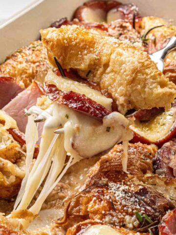 A close-up of a baked casserole dish filled with layered croissants, ham, apple slices, and melted cheese being lifted with a serving utensil. A cup and saucer are visible in the background.