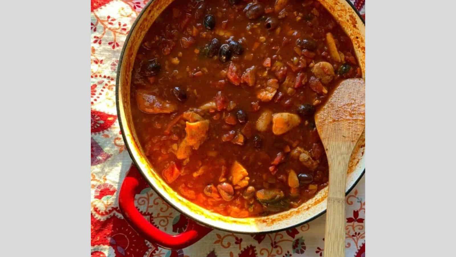 A pot of chili sits on a table with a decorative cloth, exuding the warmth of cozy bake nights. It&rsquo;s brimming with chunks of meat, beans, and tomato pieces in a rich red sauce. A wooden spoon rests inside, and the pot&rsquo;s handles are painted red, ready for your next clean plate moment.