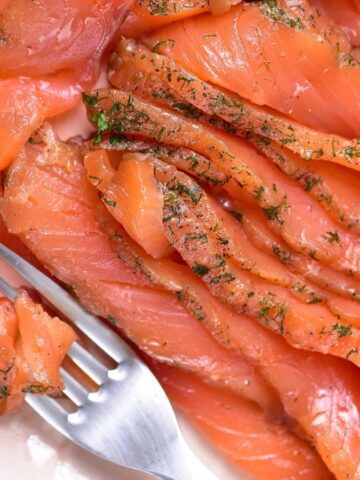 Sliced cured salmon with dill arranged on a white plate; a fork holds a small portion of the salmon.