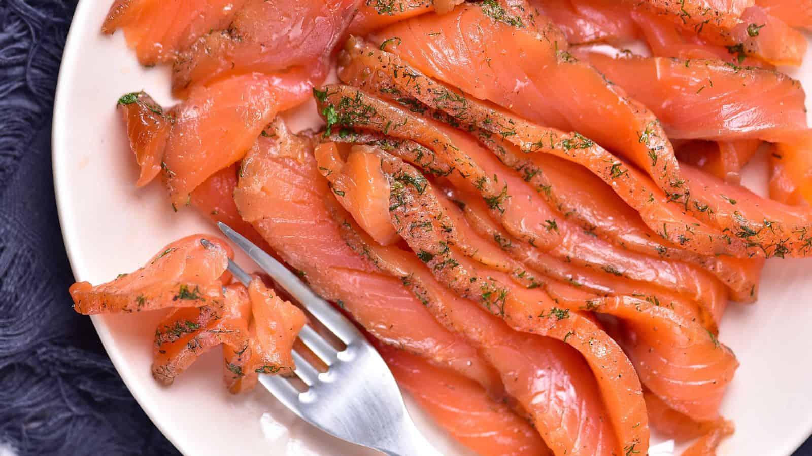 Thin slices of cured salmon with herbs are arranged on a white plate, with a fork holding a piece in the foreground.