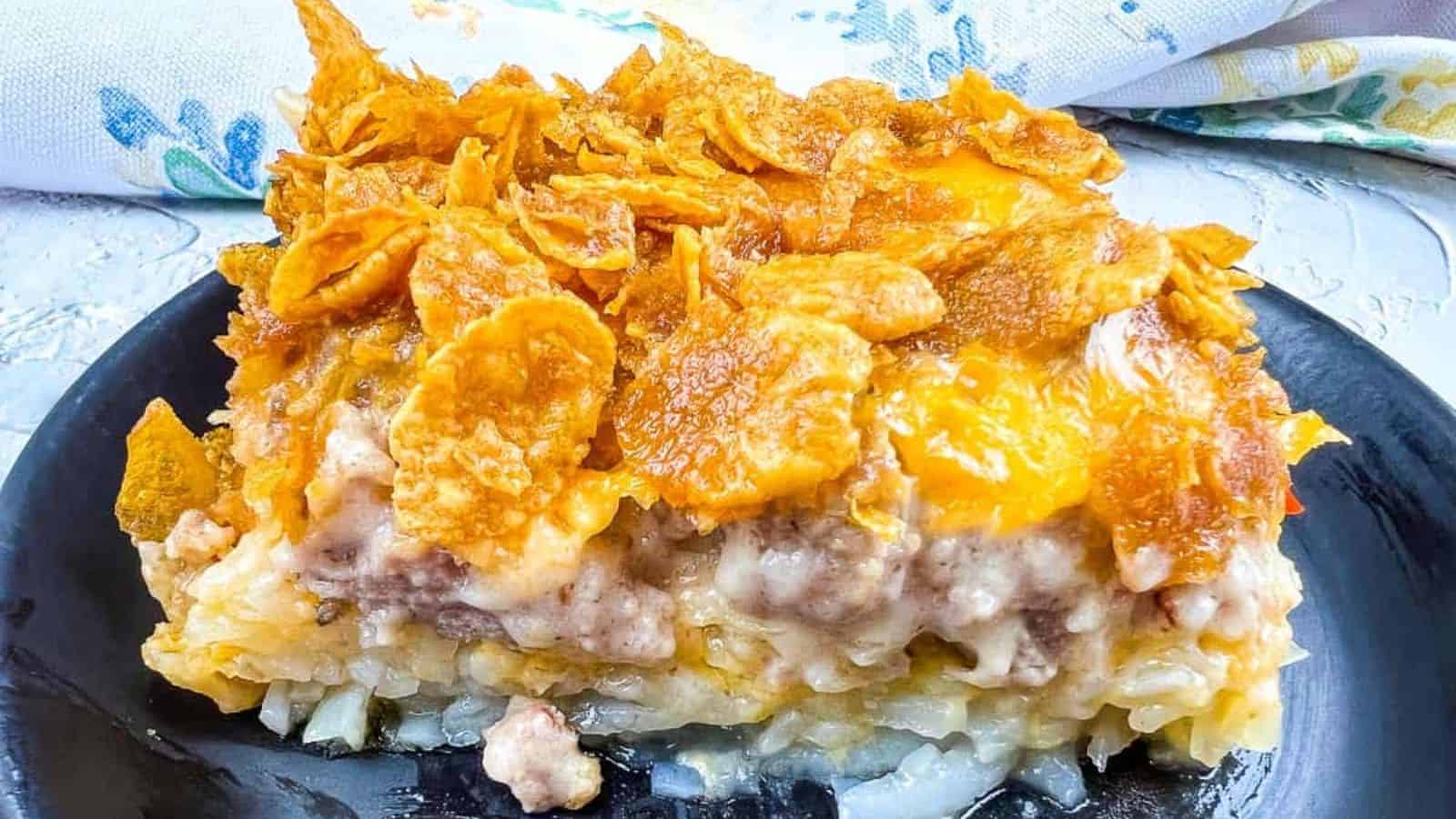 A close-up of a square serving of casserole on a black plate, showing layers of hashbrowns, ground meat, creamy sauce, melted cheese, and crispy cornflakes&mdash;classic casseroles that fill any kitchen with good memories. A patterned cloth sits in the background.
