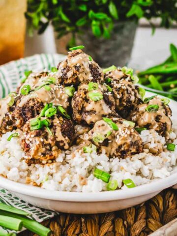 A bowl of rice topped with meatballs in a creamy sauce, garnished with chopped green onions. It is placed on a striped cloth, accompanied by a wooden spoon and a bowl of green beans. A beverage is visible in the background.