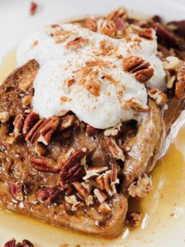 A slice of French toast on a white plate, topped with whipped cream, chopped pecans, a sprinkle of cinnamon, and drizzled with syrup.