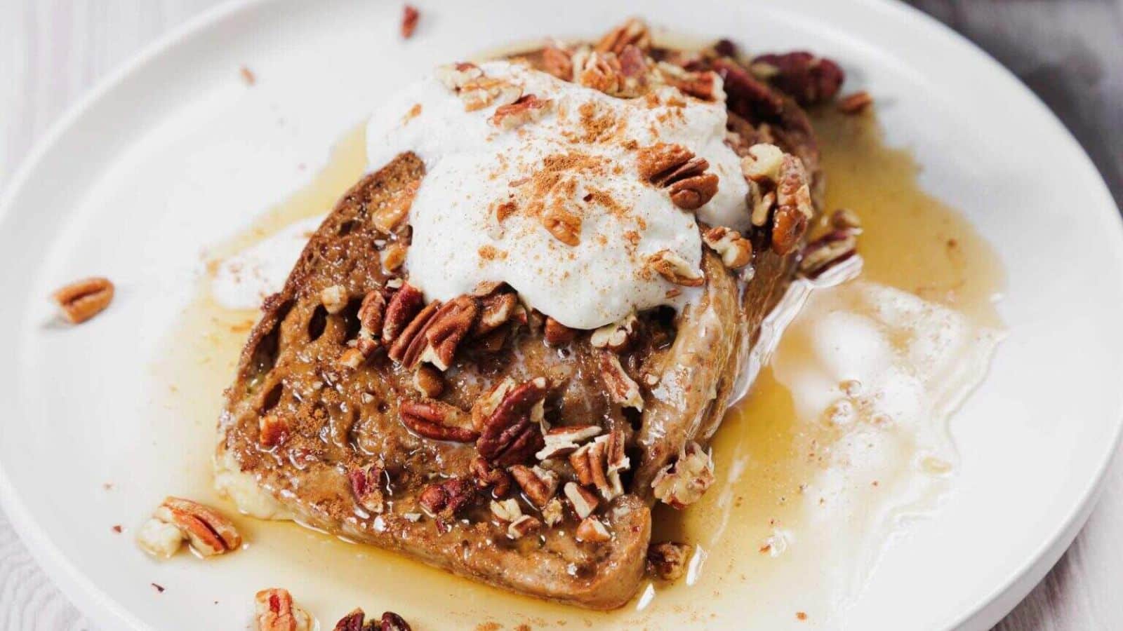 A slice of French toast on a white plate is topped with whipped cream, pecans, a dusting of cinnamon, and drizzled with syrup.
