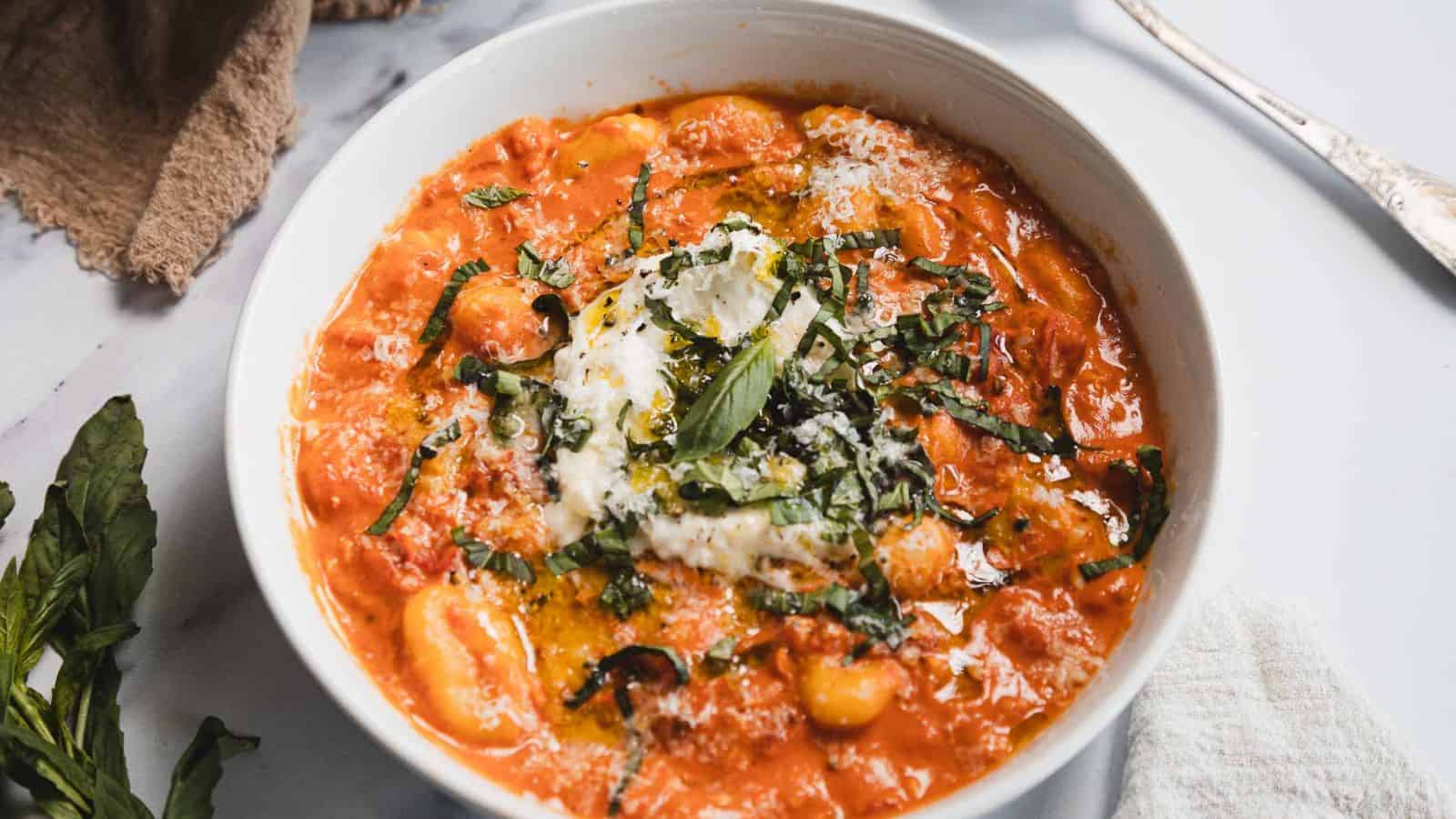 A white bowl filled with tomato-based gnocchi topped with creamy cheese, chopped fresh basil, and a drizzle of olive oil sits on a white surface with a fork, napkin, and basil leaves nearby.