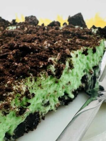 A slice of dessert with a crumbly chocolate topping and green cream filling on a white plate. A fork is placed beside the slice. The background features a yellow surface and a blurred jar.