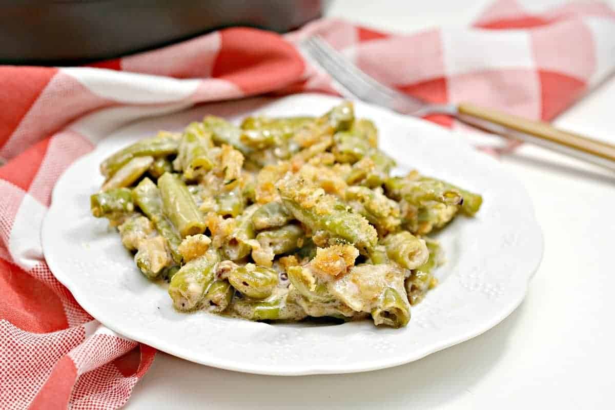 A white plate filled with creamy green bean casserole topped with breadcrumbs sits on a white surface next to a red and white checkered cloth and a fork.