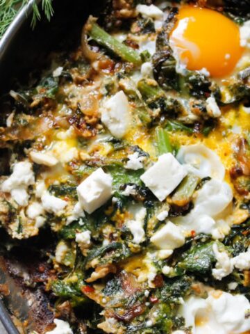 A skillet filled with a vibrant dish of cooked greens, topped with two sunny-side-up eggs and crumbled feta cheese. Fresh dill and a bowl of additional feta cubes are nearby, with a partial view of a pomegranate on the side.