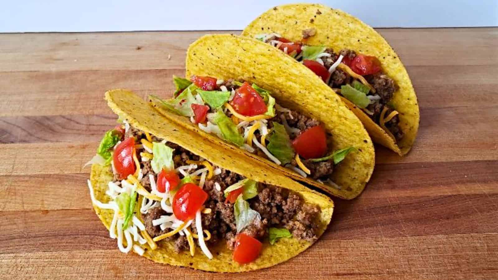 Three hard shell tacos filled with ground beef, shredded cheese, lettuce, and diced tomatoes are arranged in a row on a wooden surface&mdash;your perfect reward for getting through the week.