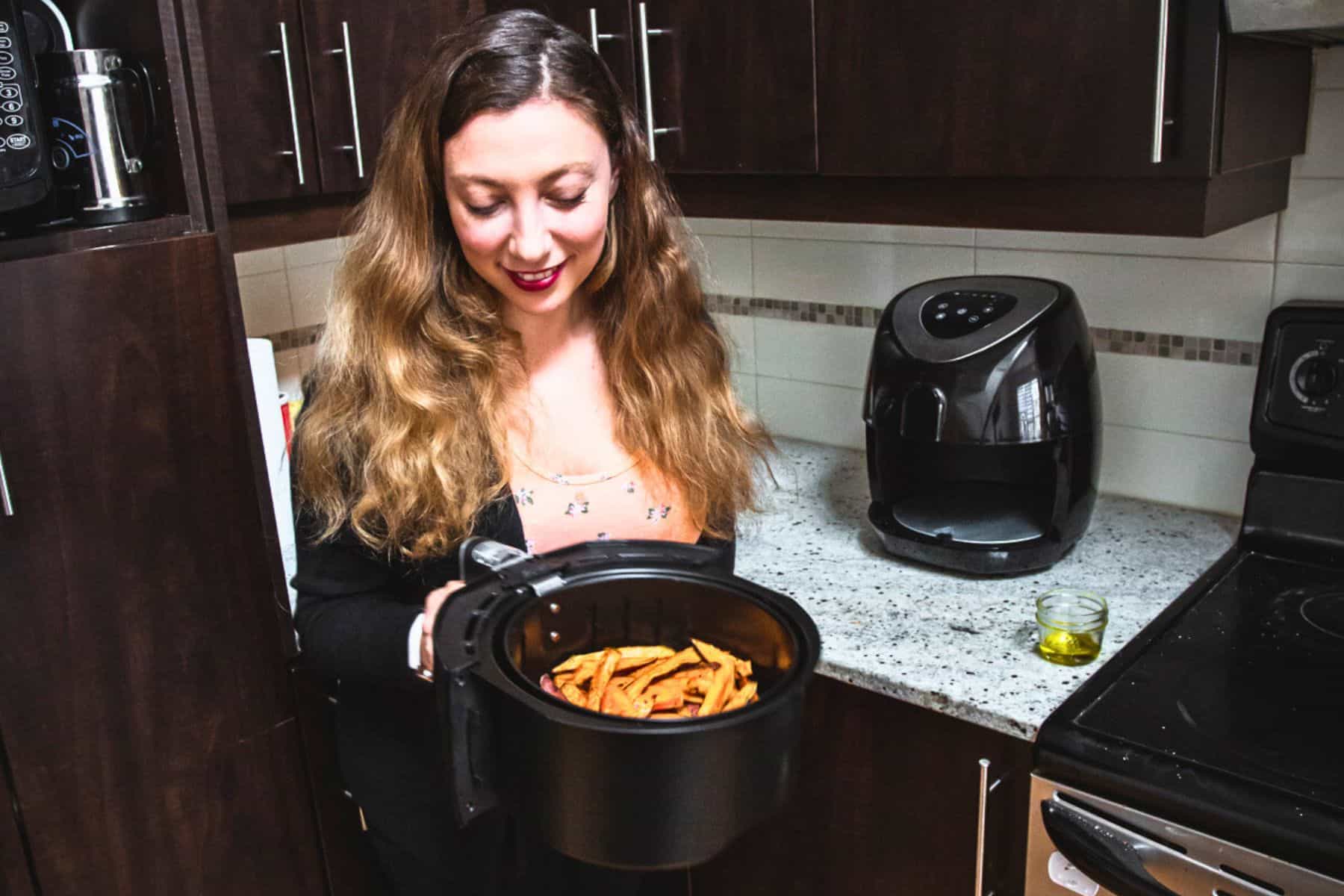 A woman with long hair stands in a kitchen, showcasing her Essential Guide to using an air fryer. She holds the basket of crispy fries, fresh from the fryer on the countertop. Nearby, a small container of oil sits by the stove, hinting at versatile cooking possibilities.