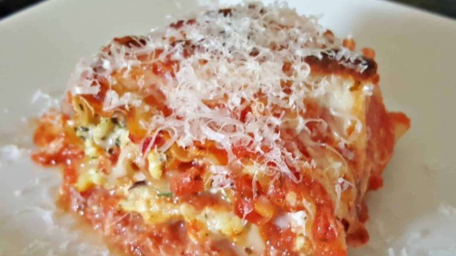A slice of lasagna topped with grated cheese on a white plate. The layers of pasta, cheese, tomato sauce, and herbs are visible, offering the perfect reward after a busy week. The dish appears freshly baked with a golden, crispy top layer&mdash;a weekend recipe delight!.