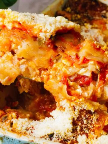 Close-up of a baked pasta dish being served from a ceramic dish. The pasta is layered with tomato sauce, cheese, and a breadcrumb topping. Partially visible basil leaves are in the background.
