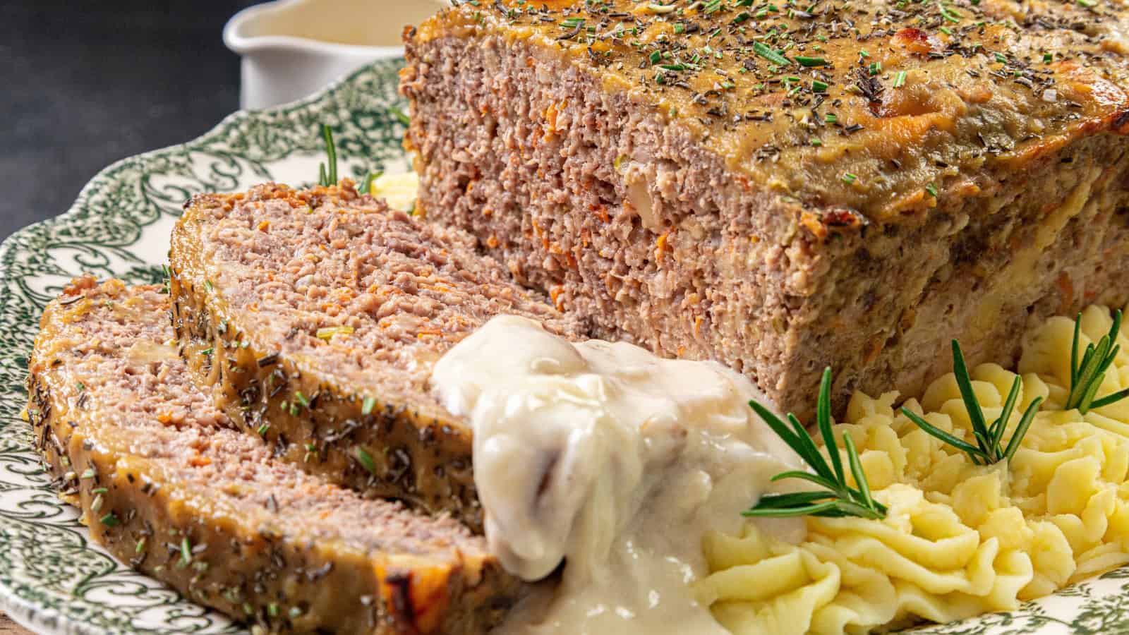 Sliced meatloaf with gravy on decorative plate, garnished with rosemary, accompanied by mashed potatoes.