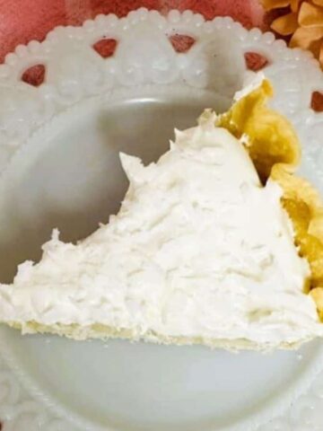 A slice of cream pie with a golden crust sits on a white decorative plate atop a pink and white striped cloth, with a light-colored flower nearby.