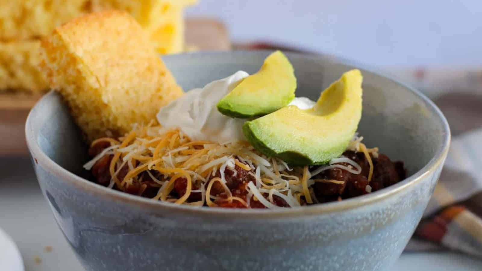 A bowl of chili crowned with shredded cheese, a dollop of sour cream, and two avocado slices sits ready for the perfect weekend meal. A piece of cornbread rests beside it on a wooden cutting board backdrop, with a white and yellow cloth adding a cozy touch to this no-plans recipe.