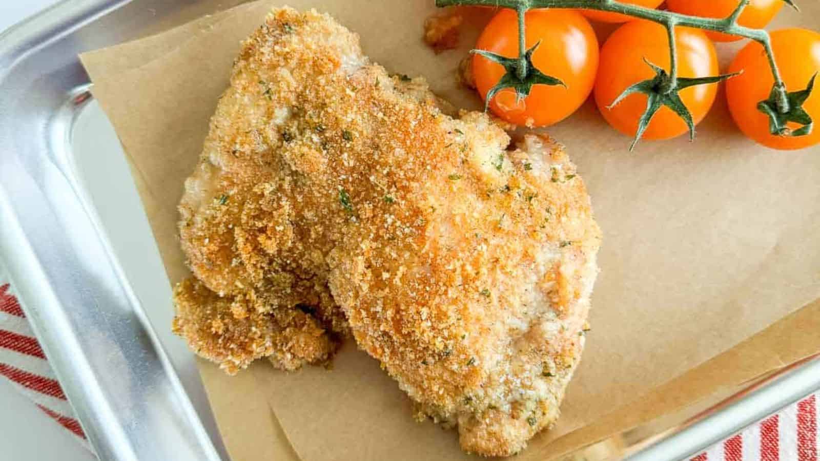 Golden brown breaded chicken cutlets rest on a parchment-lined tray, next to vine-ripened cherry tomatoes. Perfectly seasoned with herbs, this cozy bake completes the scene with a glimpse of a red and white striped cloth in the corner. A feast worthy of any clean plate!.